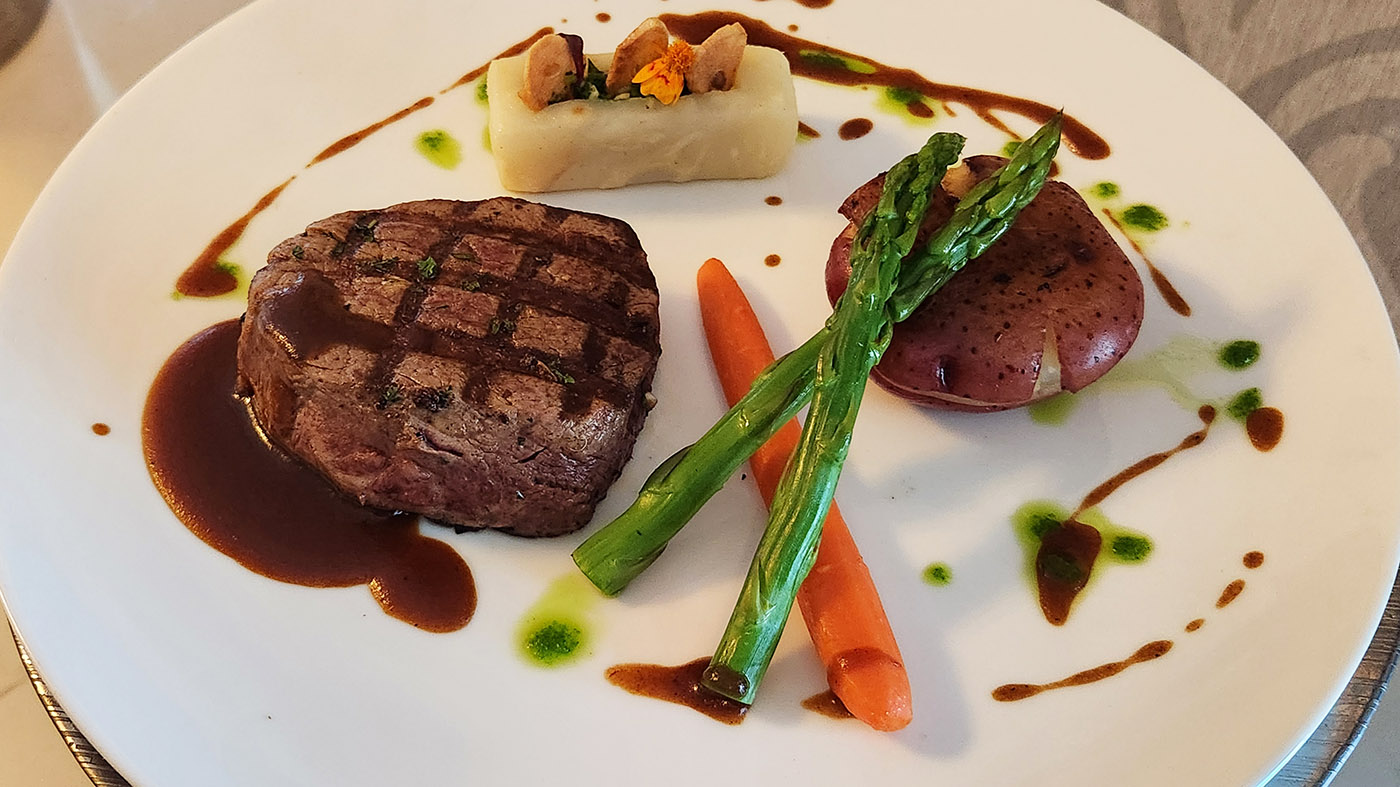 Grilled filet mignon with brown sauce, asparagus, carrot, purple potato, and a nut-topped white potato roulade on a white plate, artworked with greens and drizzles.