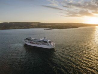 White cruise ship sailing on calm sea at sunset, coastline visible in the distance.