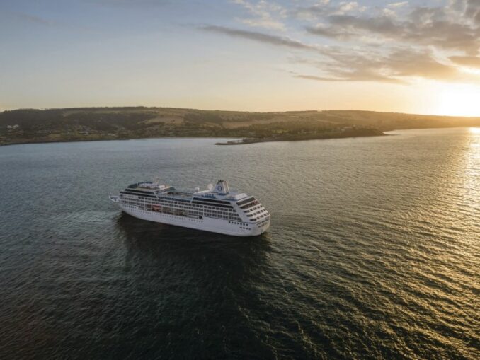 White cruise ship sailing on calm sea at sunset, coastline visible in the distance.