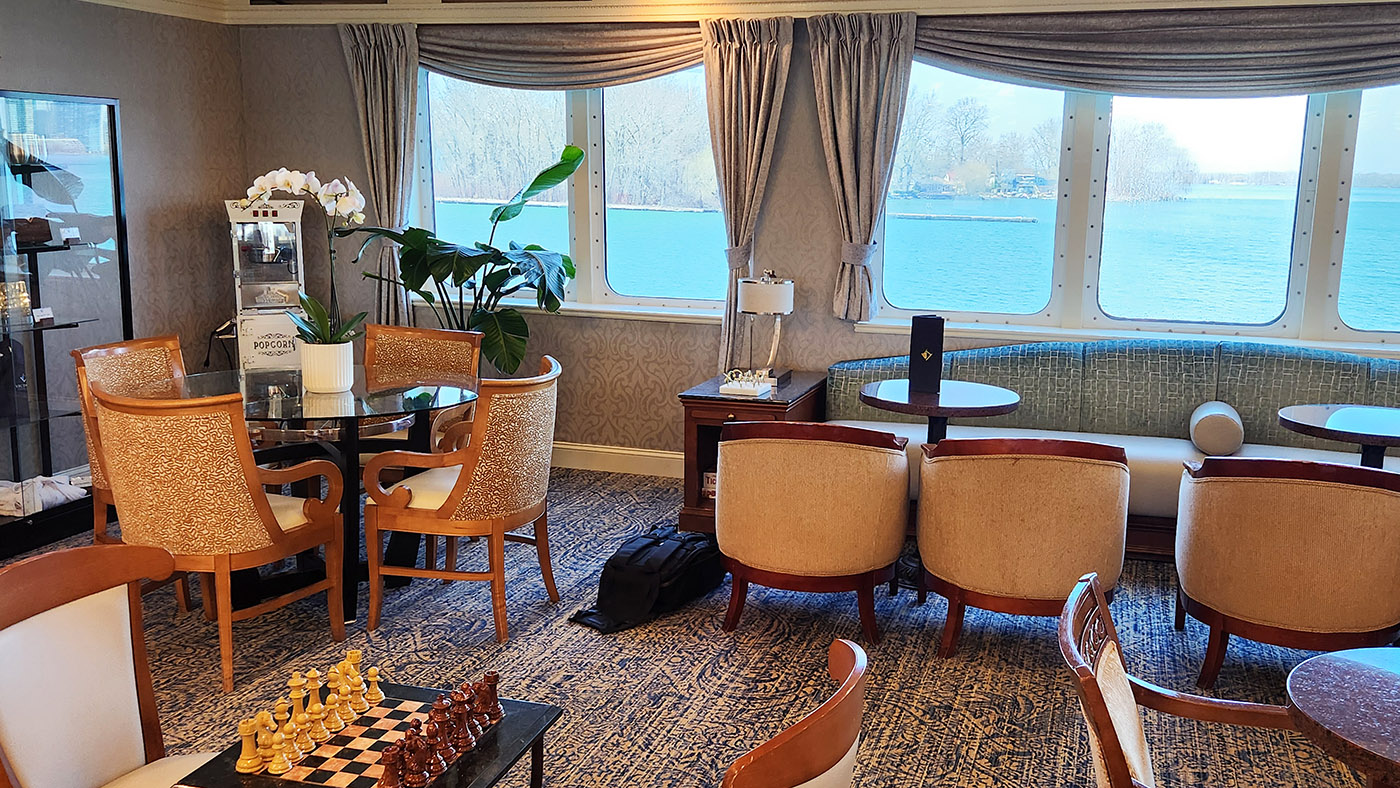 Boutique lounge on a ship with beige chairs around a glass table, a chessboard in the foreground, and large windows overlooking blue water.