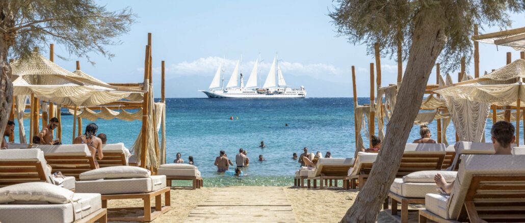 Beach club with loungers and shaded cabanas facing clear turquoise sea; a large white sailboat on the horizon.