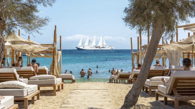 Beach club with loungers and shaded cabanas facing clear turquoise sea; a large white sailboat on the horizon.