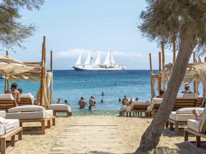Beach club with loungers and shaded cabanas facing clear turquoise sea; a large white sailboat on the horizon.