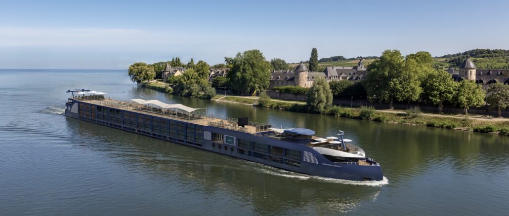 A large blue river cruise ship gliding on calm water past a green riverside town with stone buildings and trees in the background.