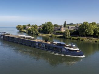 A large blue river cruise ship gliding on calm water past a green riverside town with stone buildings and trees in the background.
