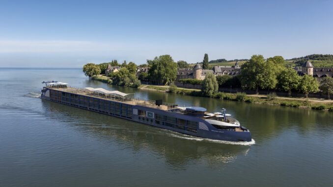 A large blue river cruise ship gliding on calm water past a green riverside town with stone buildings and trees in the background.