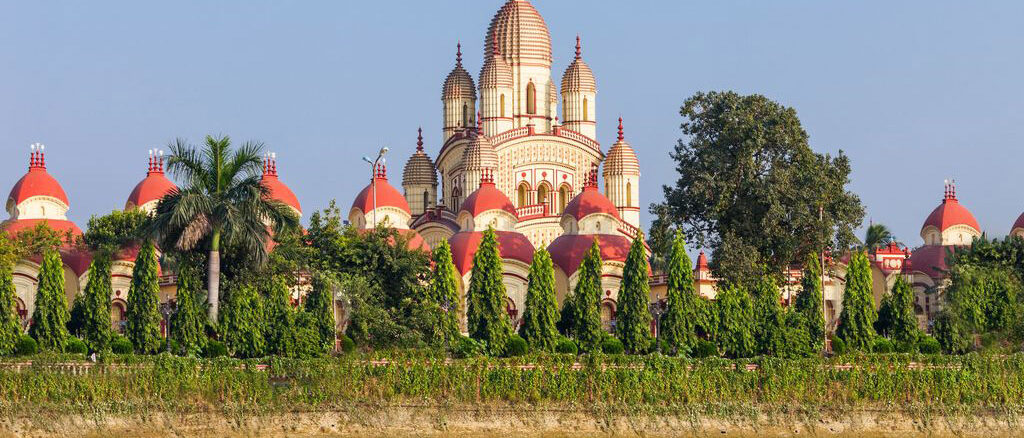 Large temple with beige and red domes visible behind a row of evergreen trees along a river front surface.