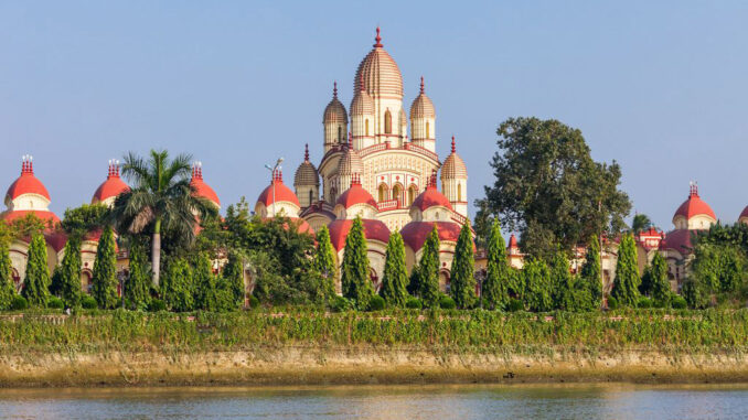 Large temple with beige and red domes visible behind a row of evergreen trees along a river front surface.