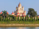 Large temple with beige and red domes visible behind a row of evergreen trees along a river front surface.