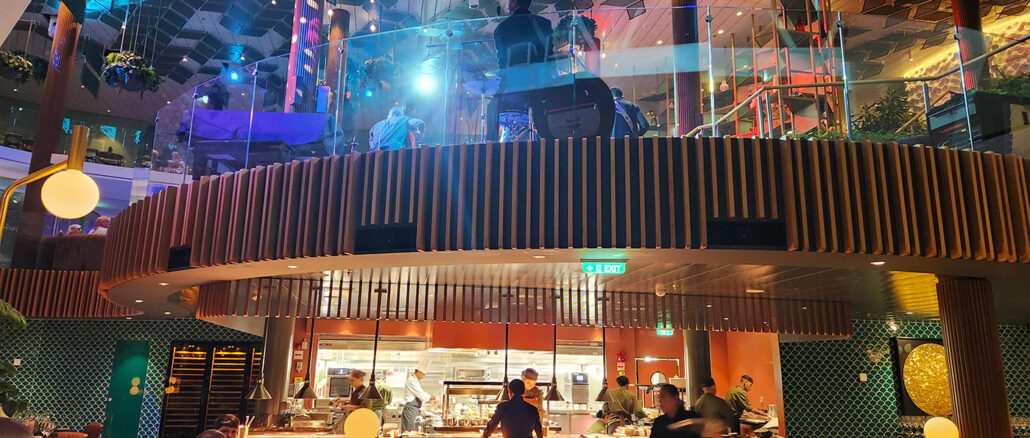 Open kitchen in a two-level restaurant with chefs at work, curved wooden balcony and colorful ambient lighting.