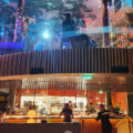Open kitchen in a two-level restaurant with chefs at work, curved wooden balcony and colorful ambient lighting.