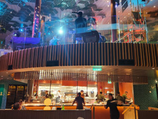 Open kitchen in a two-level restaurant with chefs at work, curved wooden balcony and colorful ambient lighting.