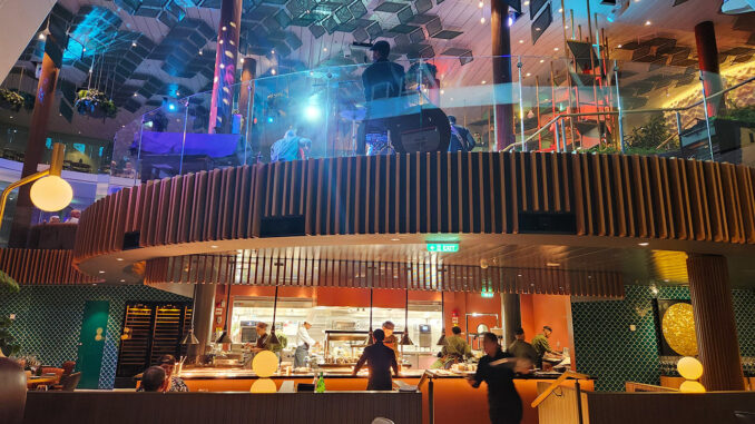 Open kitchen in a two-level restaurant with chefs at work, curved wooden balcony and colorful ambient lighting.