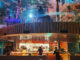 Open kitchen in a two-level restaurant with chefs at work, curved wooden balcony and colorful ambient lighting.