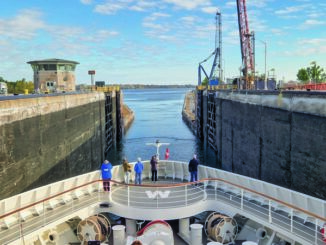 Cruise ship entering a canal lock, with tall concrete walls and cranes on the sides under a blue sky.