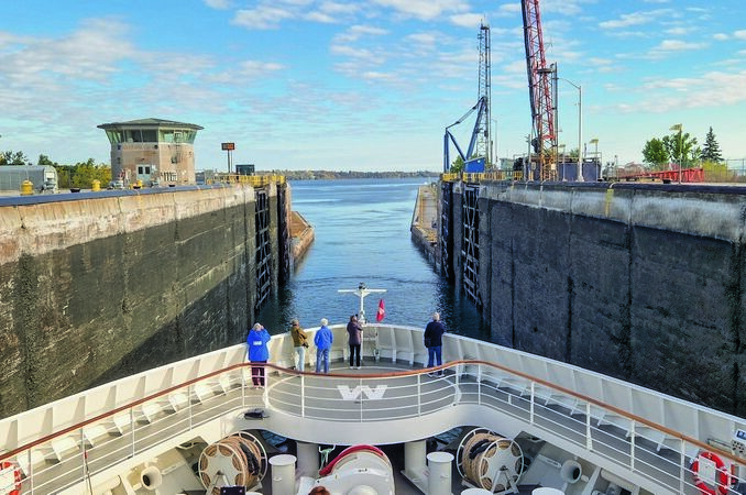 Cruise ship entering a canal lock, with tall concrete walls and cranes on the sides under a blue sky.
