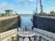 Cruise ship entering a canal lock, with tall concrete walls and cranes on the sides under a blue sky.