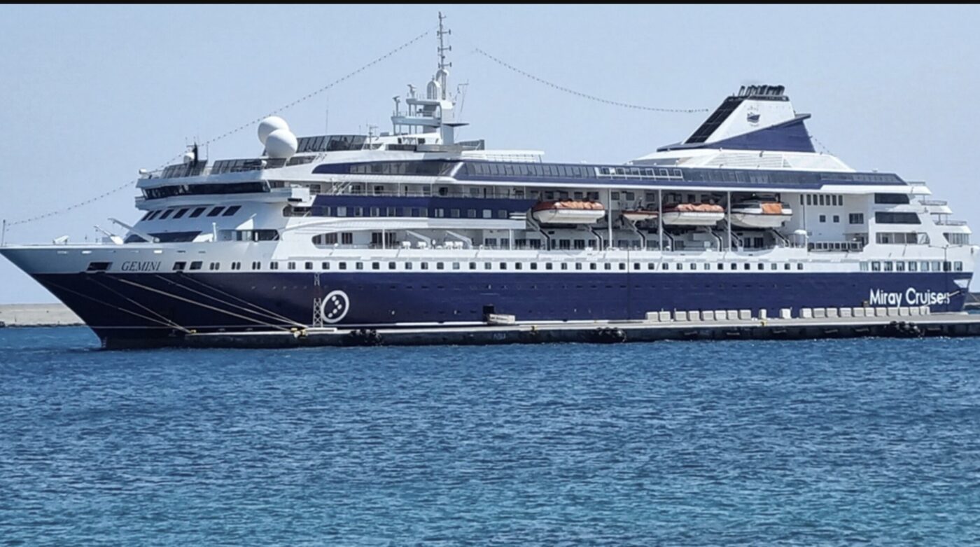Large white and navy cruise ship docked at a port, with orange lifeboats along its side.