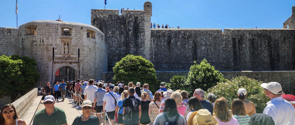 Crowd of tourists walking toward a historic stone fortress gate on a bright blue day.
