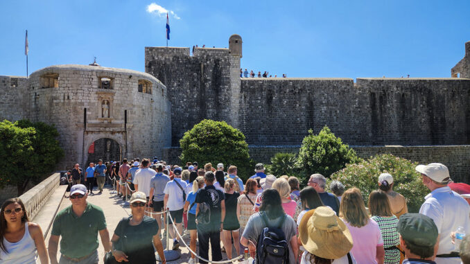 Crowd of tourists walking toward a historic stone fortress gate on a bright blue day.