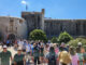 Crowd of tourists walking toward a historic stone fortress gate on a bright blue day.