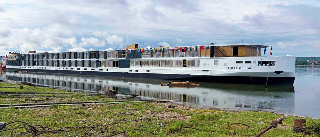 Long white river cruise ship named Emerald Lumi docked at the shore with colorful flags on the upper deck and calm water reflecting the boat.
