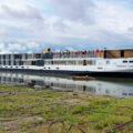 Long white river cruise ship named Emerald Lumi docked at the shore with colorful flags on the upper deck and calm water reflecting the boat.
