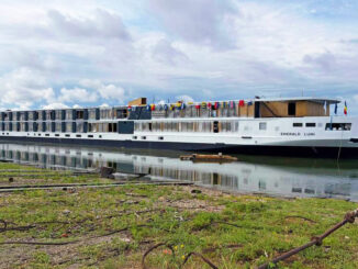 Long white river cruise ship named Emerald Lumi docked at the shore with colorful flags on the upper deck and calm water reflecting the boat.