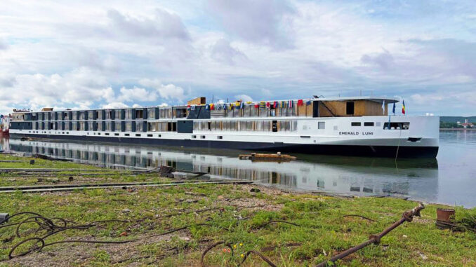 Long white river cruise ship named Emerald Lumi docked at the shore with colorful flags on the upper deck and calm water reflecting the boat.