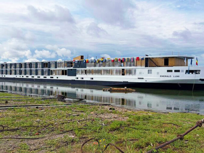 Long white river cruise ship named Emerald Lumi docked at the shore with colorful flags on the upper deck and calm water reflecting the boat.