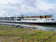 Long white river cruise ship named Emerald Lumi docked at the shore with colorful flags on the upper deck and calm water reflecting the boat.