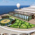 A cruise ship deck featuring a circular mini-golf course adjacent to a glass-walled lounge and a pool, with the ocean in the background, under a blue sky.
