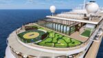 A cruise ship deck featuring a circular mini-golf course adjacent to a glass-walled lounge and a pool, with the ocean in the background, under a blue sky.