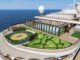 A cruise ship deck featuring a circular mini-golf course adjacent to a glass-walled lounge and a pool, with the ocean in the background, under a blue sky.