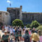 Crowd of tourists walking toward a historic stone fortress gate on a bright blue day.