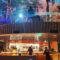 Open kitchen in a two-level restaurant with chefs at work, curved wooden balcony and colorful ambient lighting.