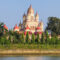 Large temple with beige and red domes visible behind a row of evergreen trees along a river front surface.