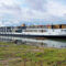 Long white river cruise ship named Emerald Lumi docked at the shore with colorful flags on the upper deck and calm water reflecting the boat.